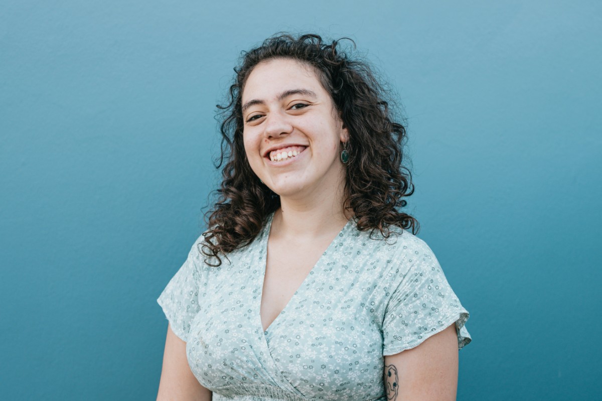 Person with curly hair smiling in front of a blue background, wearing a light floral dress.