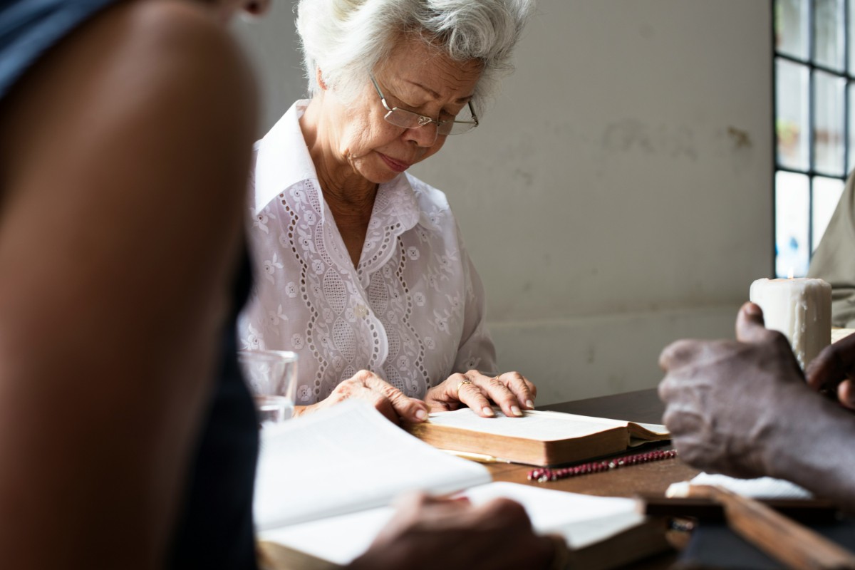 Older woman with gray hair reading a book at a table, accompanied by others, in a bright room.