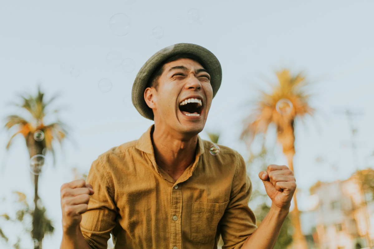 Person wearing a hat and yellow shirt, joyfully raising fists with a backdrop of palm trees and a clear sky.