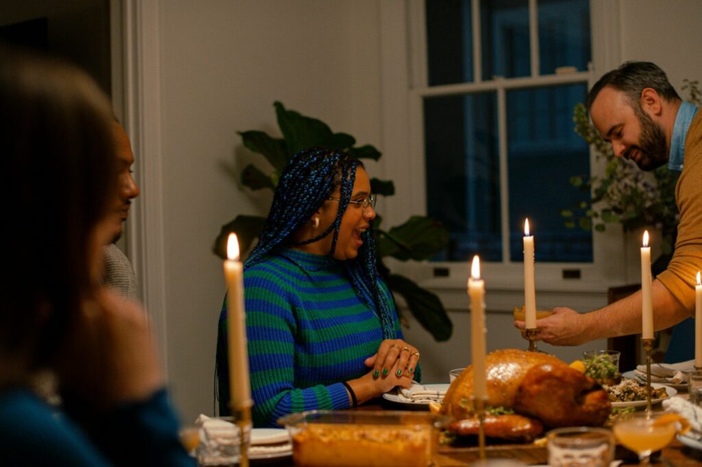 A group of people sitting at a dinner table with lit candles, engaging in conversation. There are dishes of food, including a roasted turkey, in front of them.