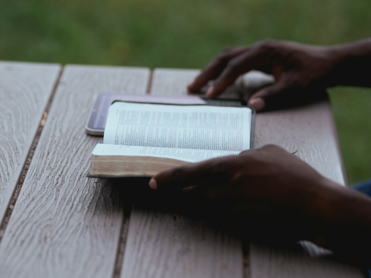 Person reading a book at an outdoor wooden table, with a smartphone beside them.