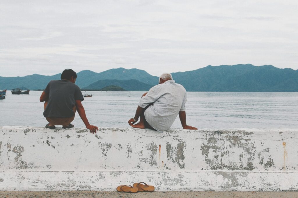 Two people sit on a concrete barrier facing a body of water with mountains in the background. One person is barefoot, with sandals on the ground.