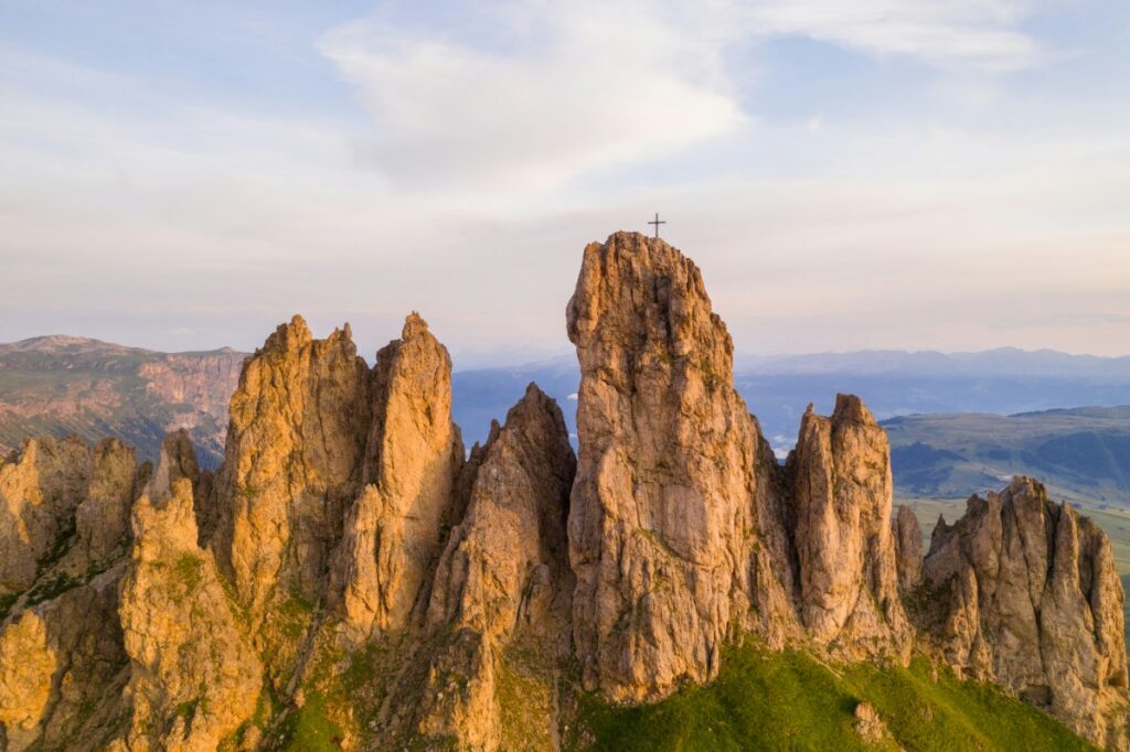 Rugged mountain peaks with a prominent central pinnacle topped by a cross, set against a cloudy sky and green landscape.