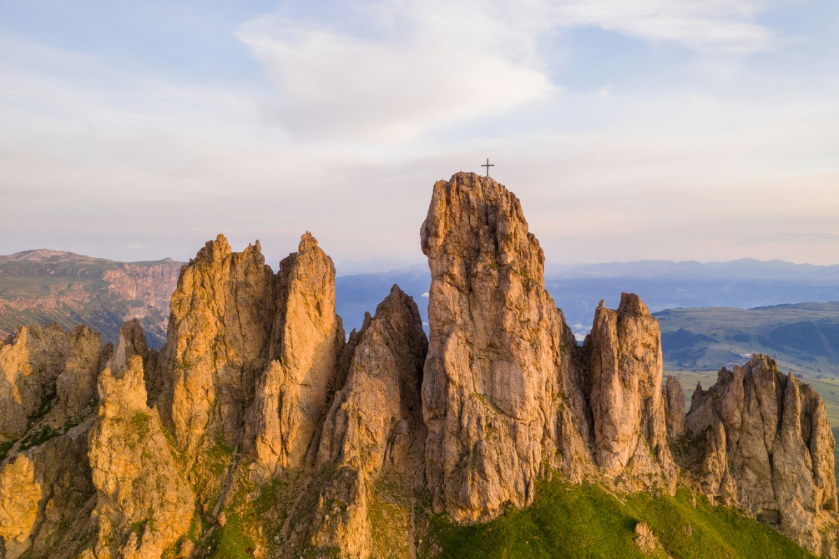Rugged mountain peaks with a prominent central pinnacle topped by a cross, set against a cloudy sky and green landscape.