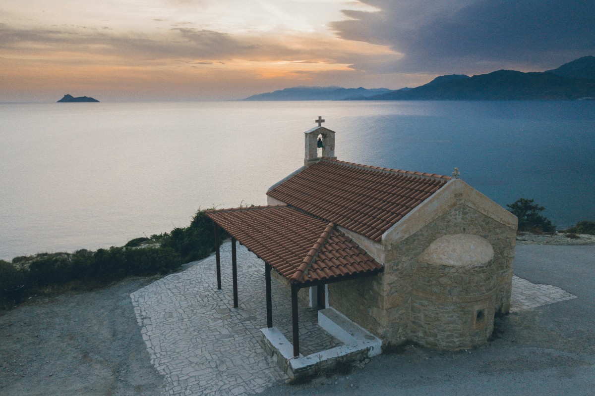 A small stone chapel with a red-tiled roof overlooking a calm body of water at sunset, with hills in the background.