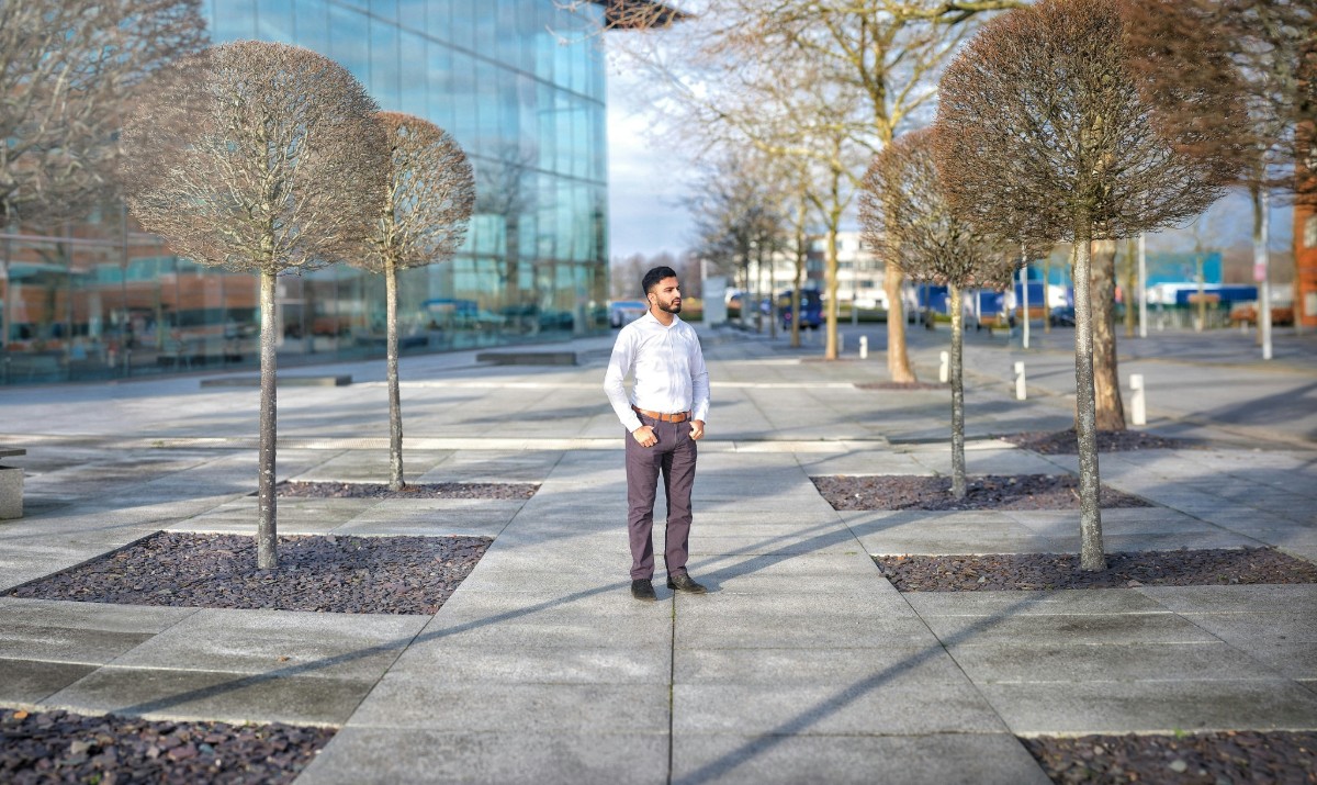 A man stands on a concrete path lined with pruned trees, near a modern glass building on a clear day.