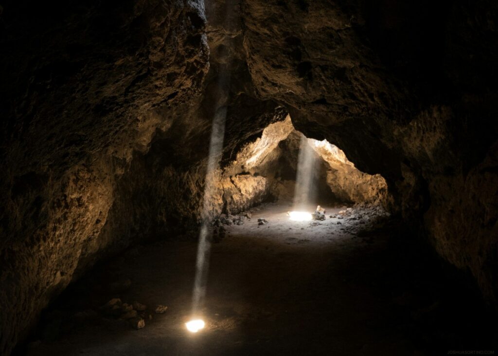 A dimly lit cave interior with two beams of light shining through openings in the ceiling onto the rocky floor.