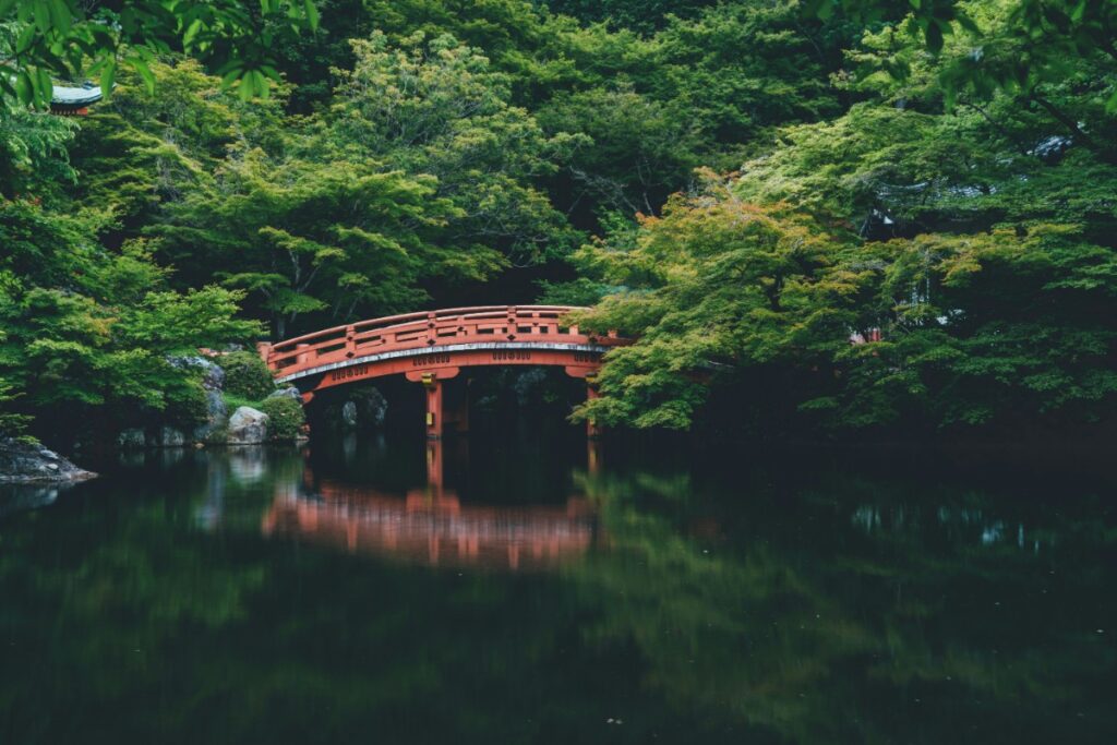 A red arched wooden bridge spans over a calm pond surrounded by lush green trees.