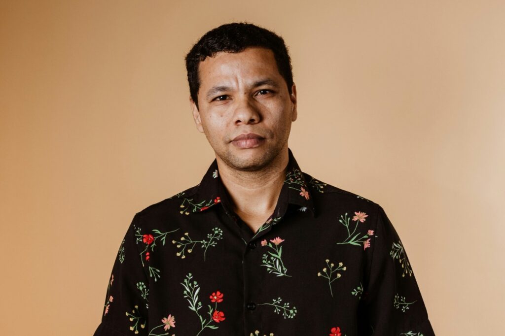 A person wearing a black floral shirt stands against a beige background.