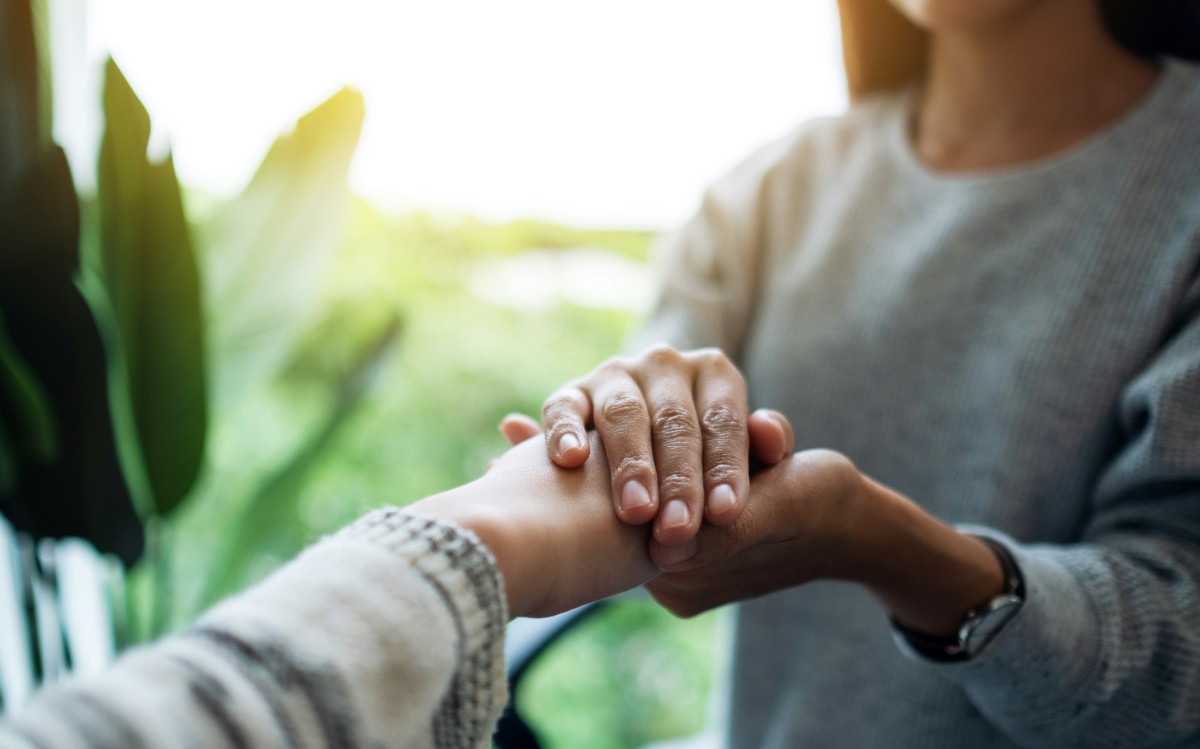 Two people are holding hands in a comforting gesture, with a blurred green background.