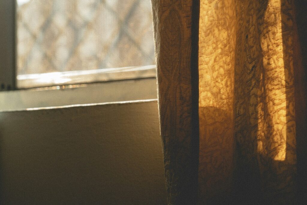 Golden sunlight filters through a textured curtain next to a window, casting soft shadows on the wall.
