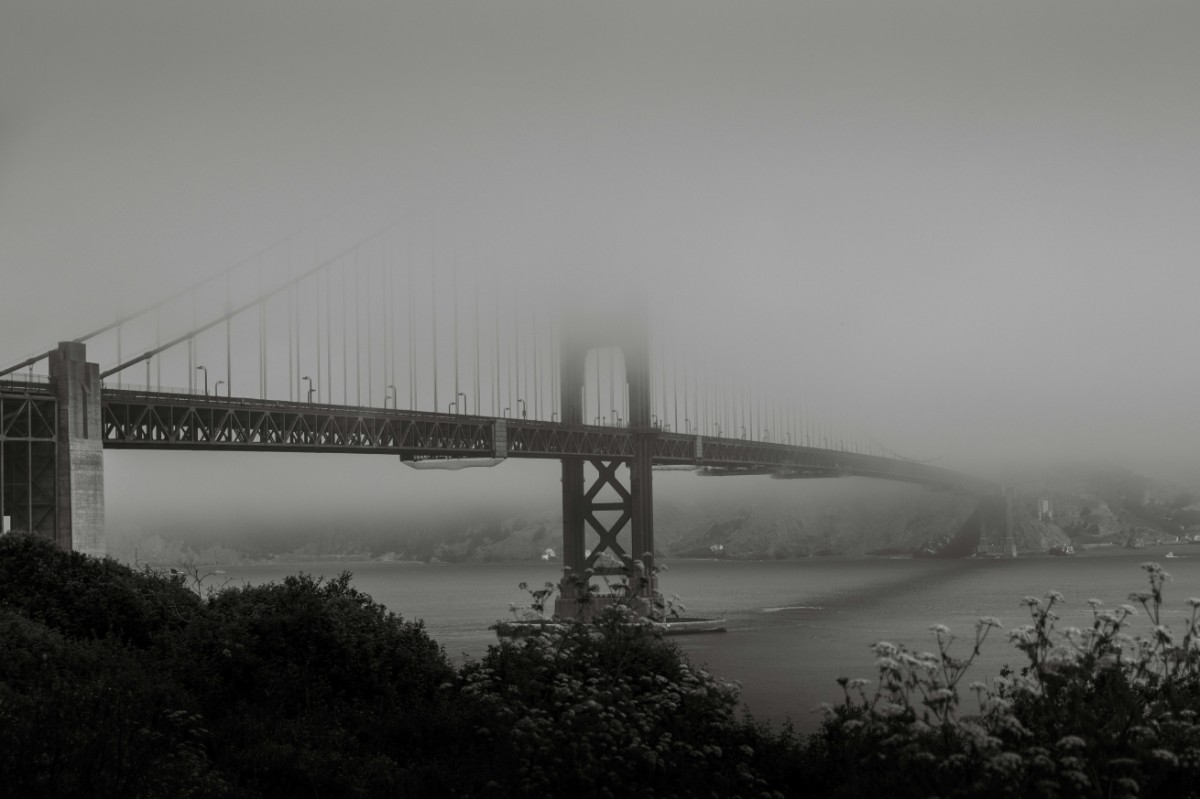 Foggy view of a large suspension bridge spanning across the water with the city barely visible in the background.