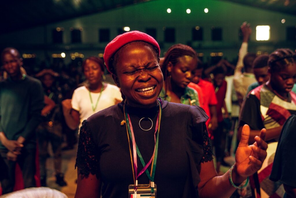 A woman wearing a red beret and lanyard passionately sings or prays in a crowded indoor gathering. Others around her are similarly engaged.