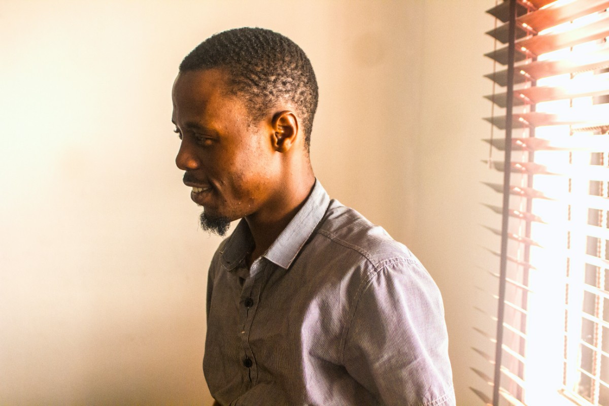 A man in a button-up shirt stands indoors near window blinds.