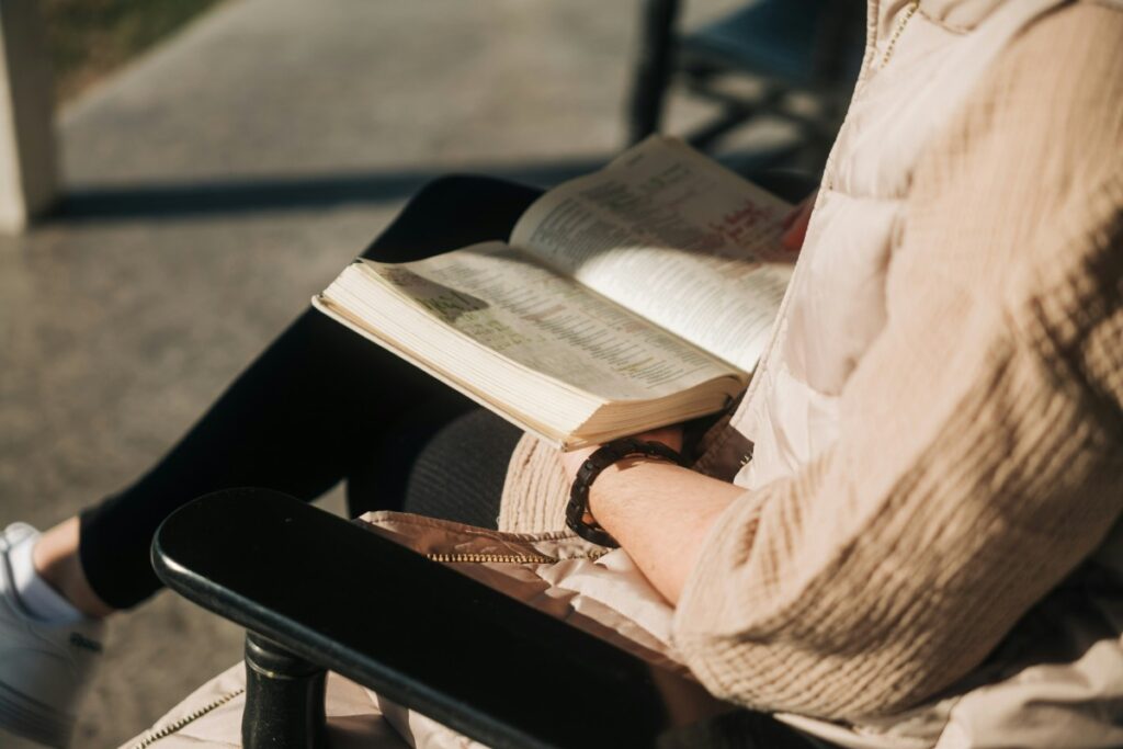 Person sitting outdoors, wearing a beige shirt, is reading an open book on their lap.