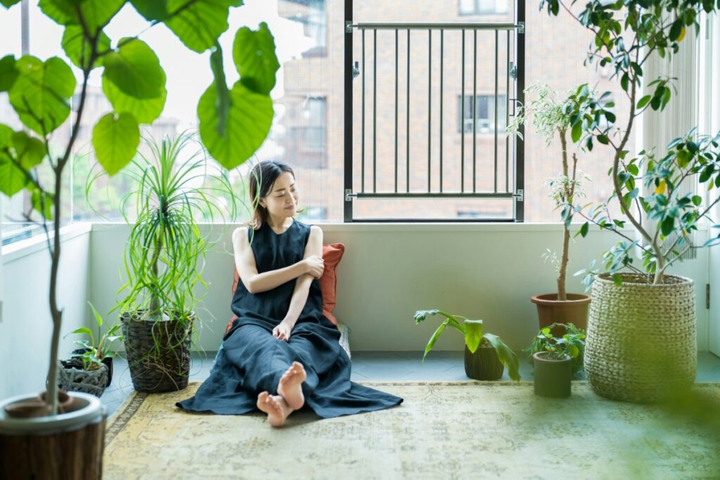 Woman sitting barefoot on the floor near a window, surrounded by various potted plants.