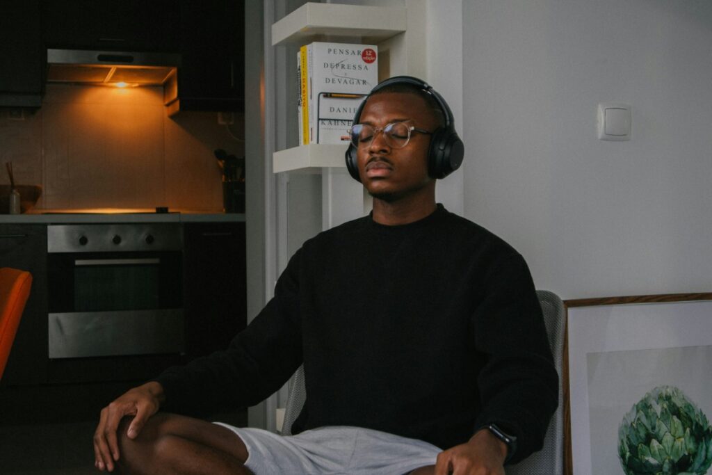 A person wearing headphones sits cross-legged in a room, eyes closed, appearing relaxed. A kitchen and a bookshelf are visible in the background.