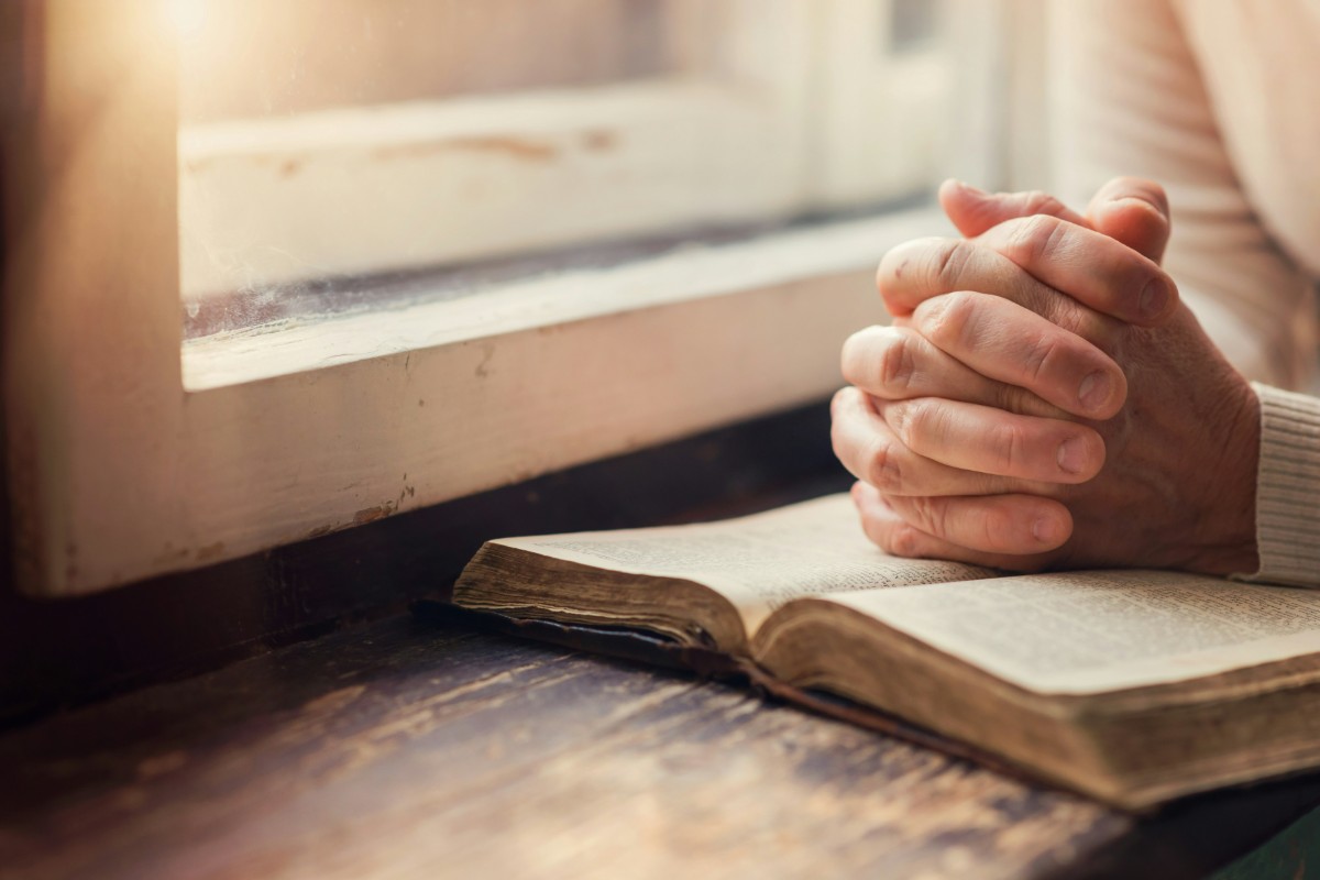 Hands clasped in prayer over an open book on a windowsill with sunlight streaming in.