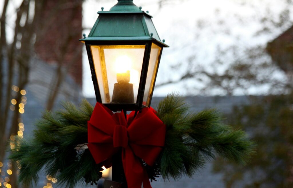 A green lantern decorated with pine branches and a red bow is lit against a blurred outdoor background.
