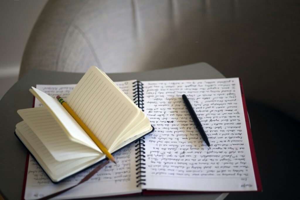 Open spiral notebook with handwritten text and a pen on a small table. A closed notebook with a pencil rests on top.