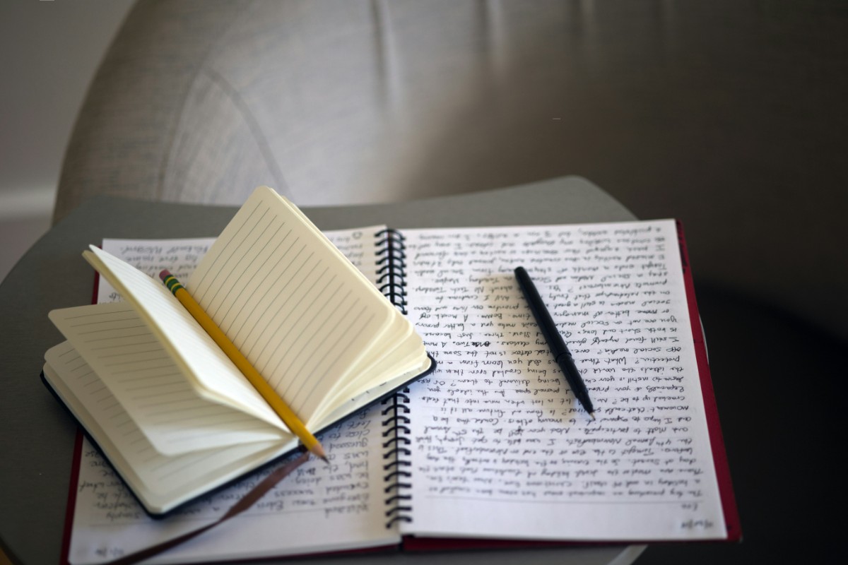 Open spiral notebook with handwritten text and a pen on a small table. A closed notebook with a pencil rests on top.