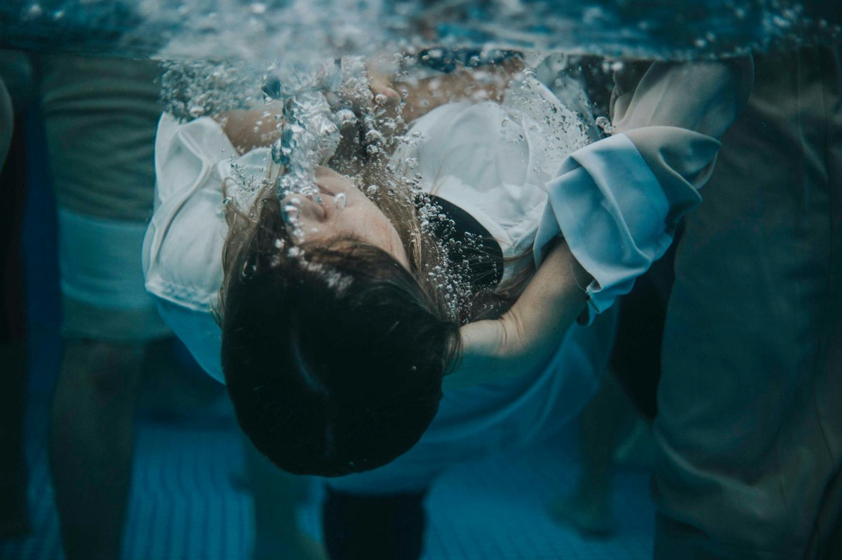 A person in a white shirt submerged underwater, surrounded by bubbles, with other people partially visible around them.
