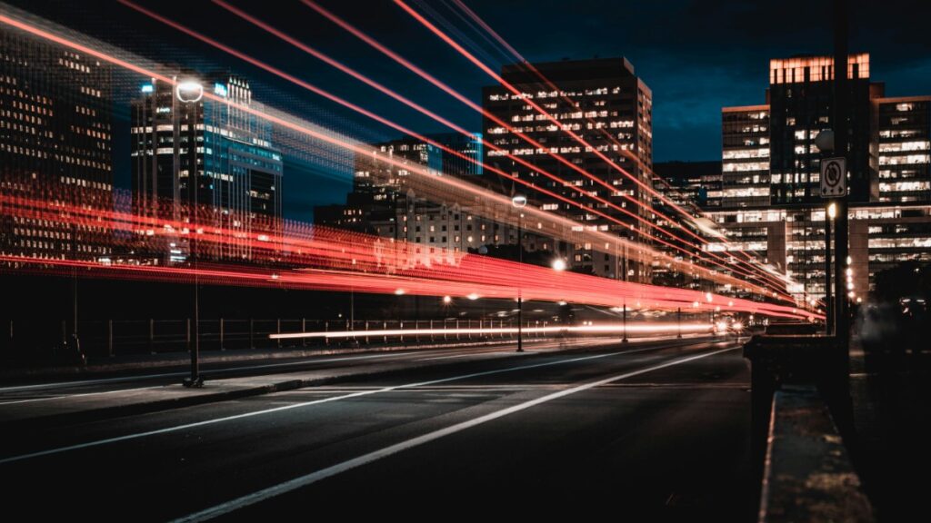 Cityscape at night with streaks of red and white light from moving vehicles, set against illuminated buildings under a dark sky.