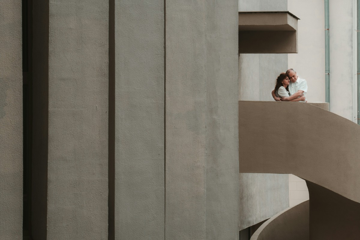 A couple embraces on a curved balcony against a backdrop of large concrete walls.
