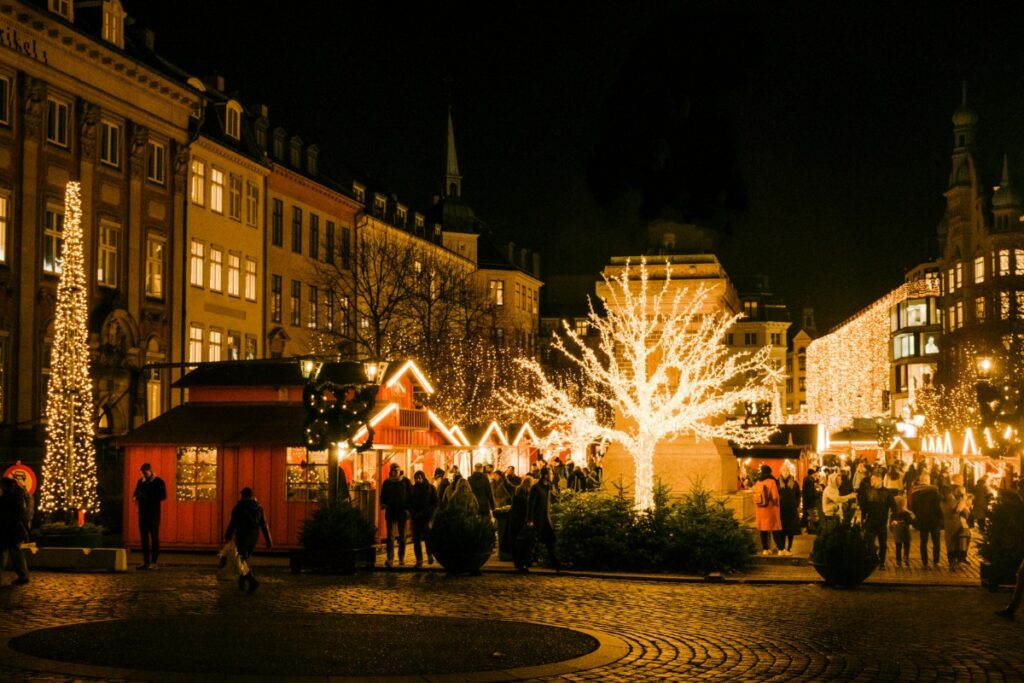 Festive Christmas market at night with illuminated trees and buildings, wooden stalls, and a crowd of people walking along a cobblestone street.