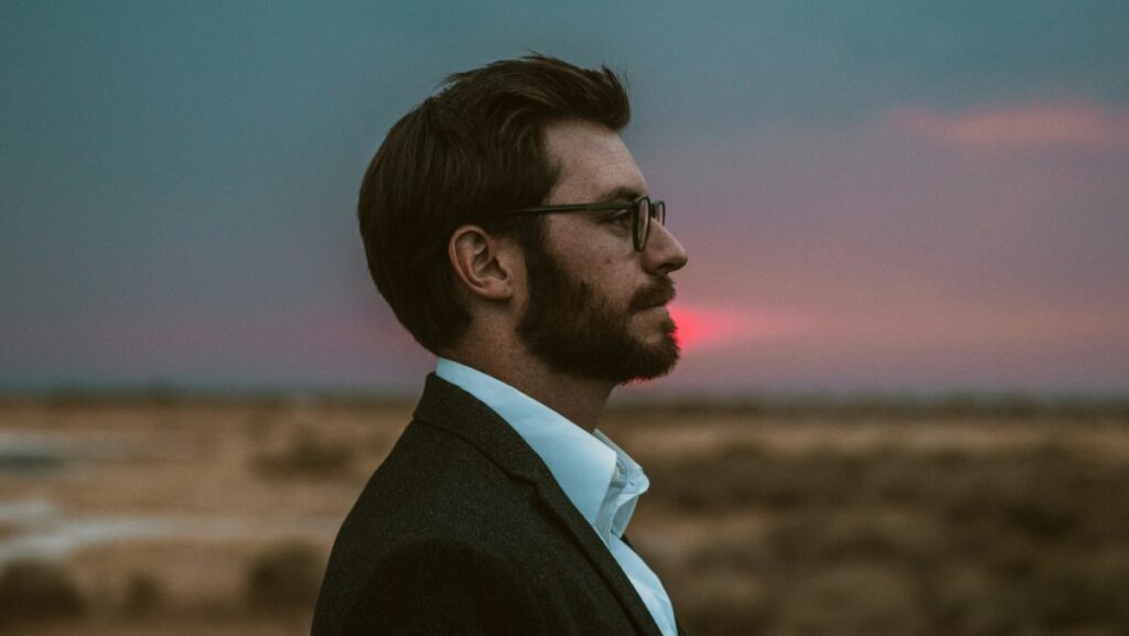 A man with glasses and a beard looks into the distance against a sunset desert backdrop.