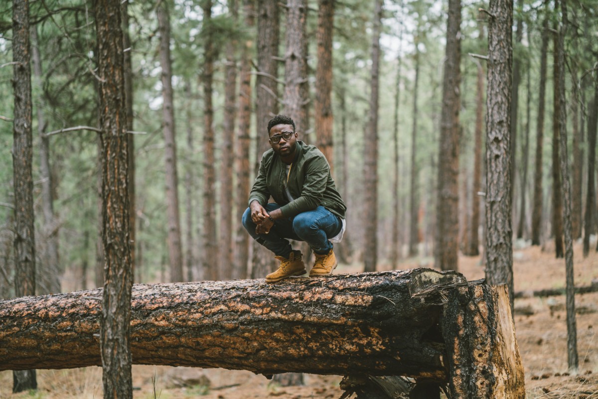 Person crouching on a fallen tree in a forest, surrounded by tall pine trees.