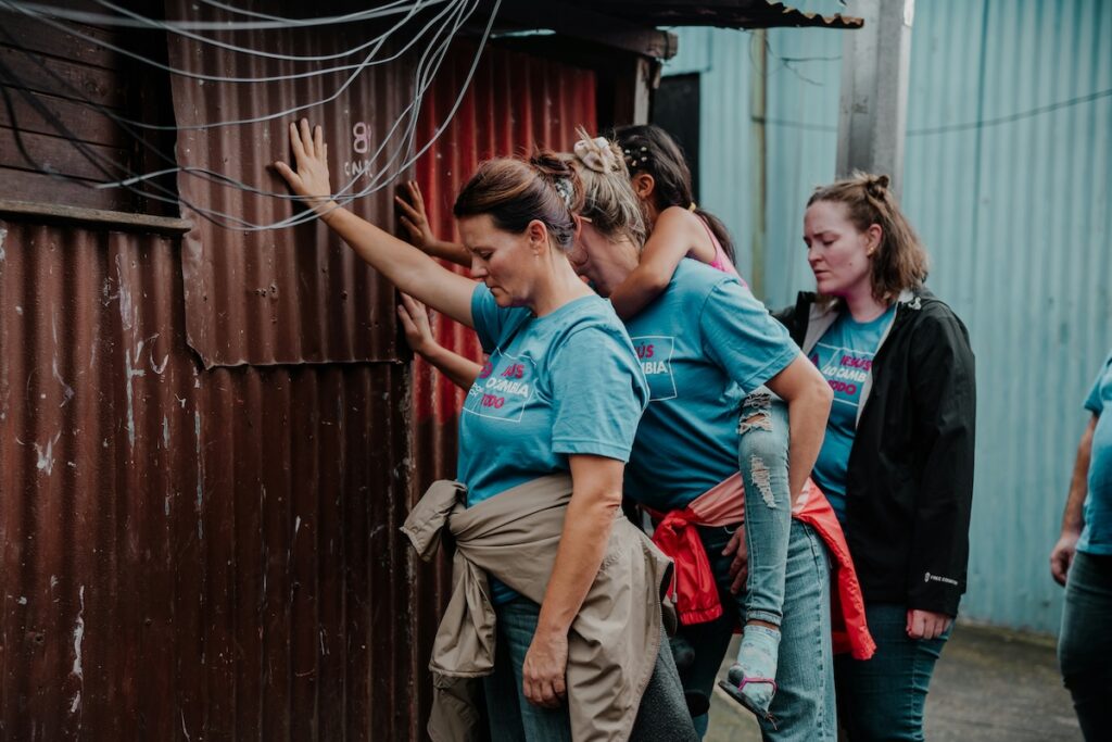 A group of women in blue shirts stands in front of a corrugated metal building. One woman is carrying a child on her back.