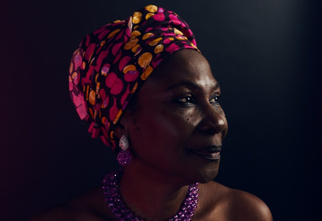 A woman wearing a colorful headwrap and purple beaded necklace and earrings, gazes to the side against a dark background.