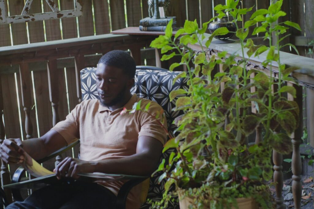 A person sitting outdoors on a patterned chair, reading a book. A potted plant is in the foreground, and a wooden fence is in the background.