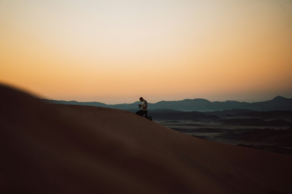 Person sits on a sand dune under a sunset sky, with distant mountains visible in the background.