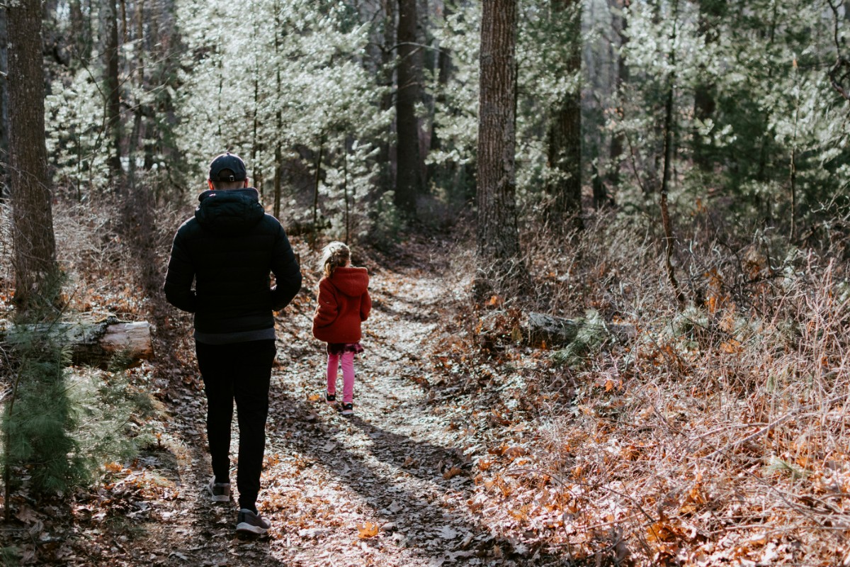 Two people walking on a forest trail, one adult in dark clothing and a child in a red coat. Trees and dry leaves surround the path.