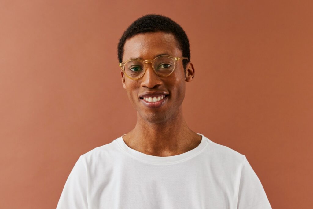 Person wearing glasses and a white t-shirt smiles against a brown background.
