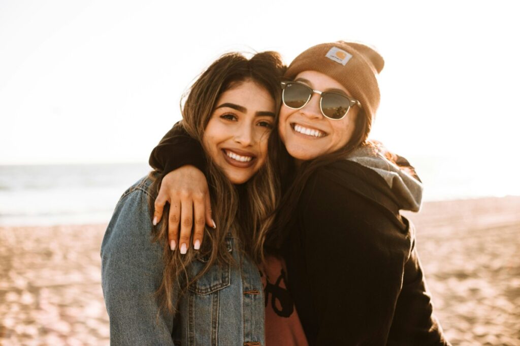 Two people smiling and embracing on a sunny beach, with one wearing a beanie and sunglasses.