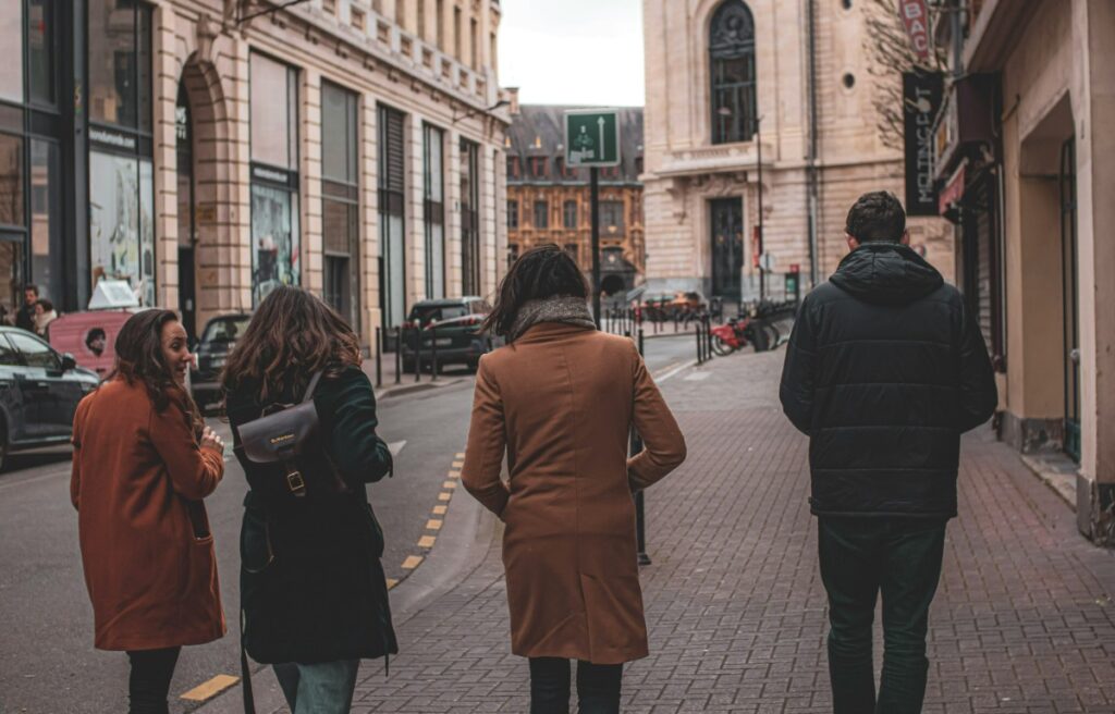 Four people walk down a narrow city street lined with tall buildings and parked cars. The road is paved with bricks, and the sky is overcast.