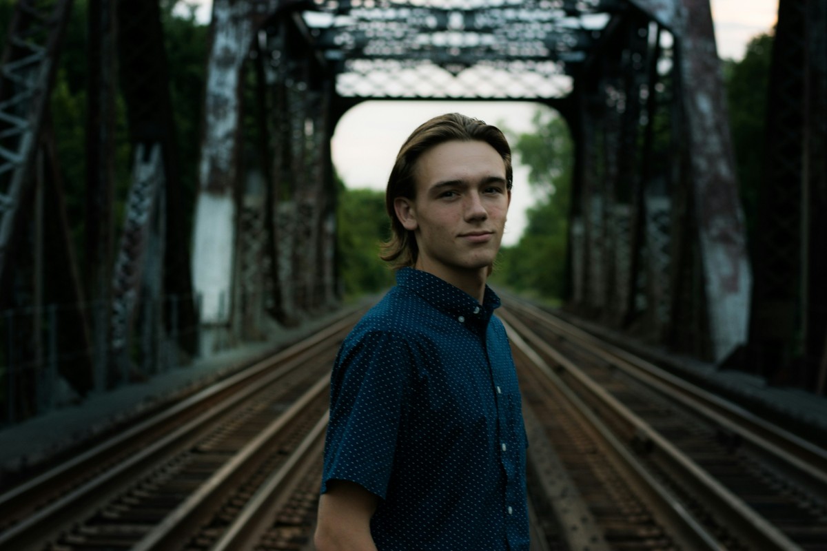 A person in a blue shirt stands on a railway bridge, facing the camera. The background shows metal bridge structure and trees.
