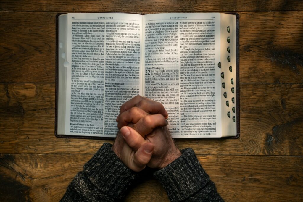 Hands clasped in prayer rest on an open Bible, which is on a wooden table.