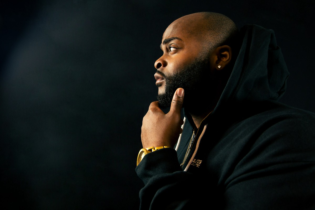 A person in a black hoodie, seen in profile, rests their chin on their hand against a dark background.