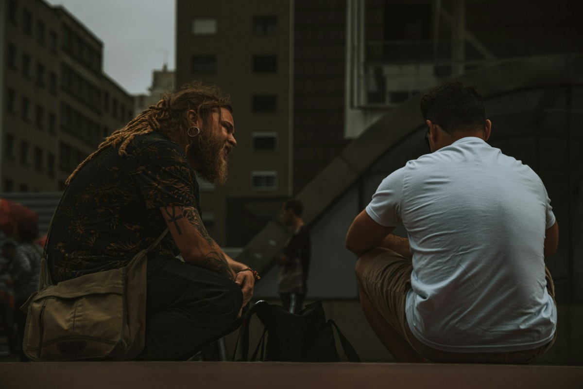 Two people are sitting outdoors on a ledge, engaged in conversation. One has long hair and a beard, while the other has short hair. Urban buildings are visible in the background.