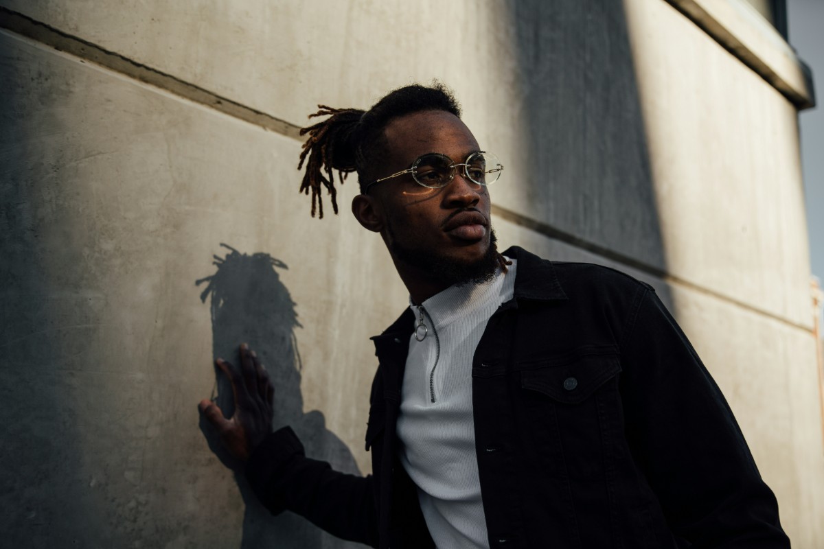 A person stands against a concrete wall, looking to the side. They have dreadlocks and wear glasses, a dark jacket, and a light shirt. Shadows cast on the wall.