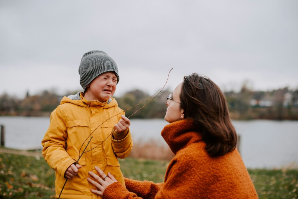 A child in a yellow coat and gray beanie appears upset while holding a stick. A woman in an orange coat kneels beside them outdoors near a body of water.