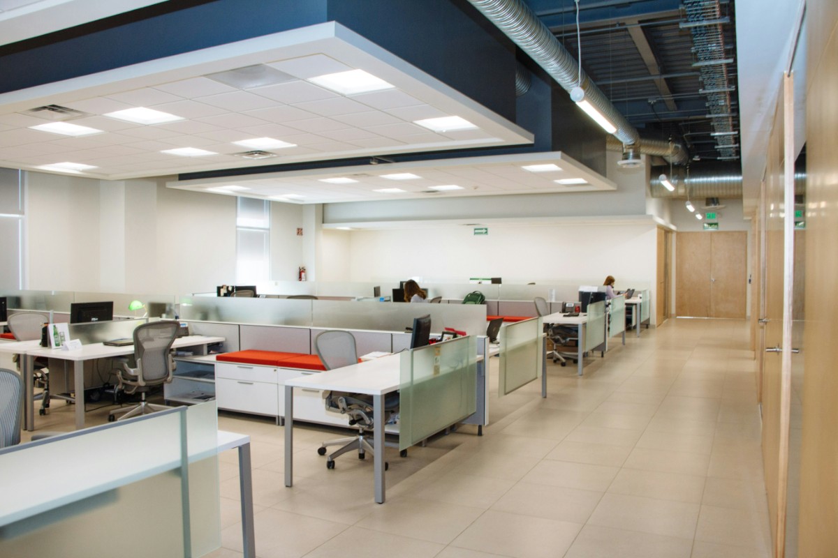 Modern office space with rows of cubicles, chairs, and desks under fluorescent lighting. Neutral colors and glass partitions are visible.