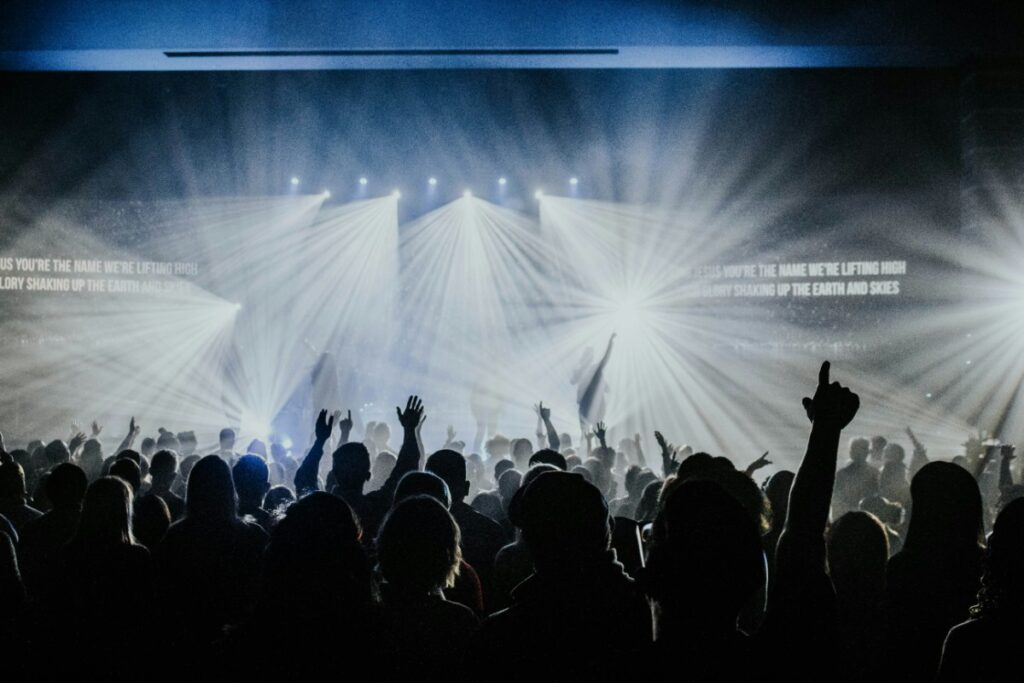 A large audience facing a brightly lit stage with beams of light and projected text. Silhouettes of people with raised hands are visible.