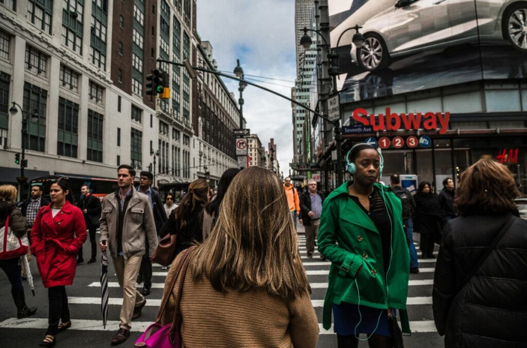 A busy city street with people crossing at a crosswalk. Buildings and an advertisement for a car are visible in the background.