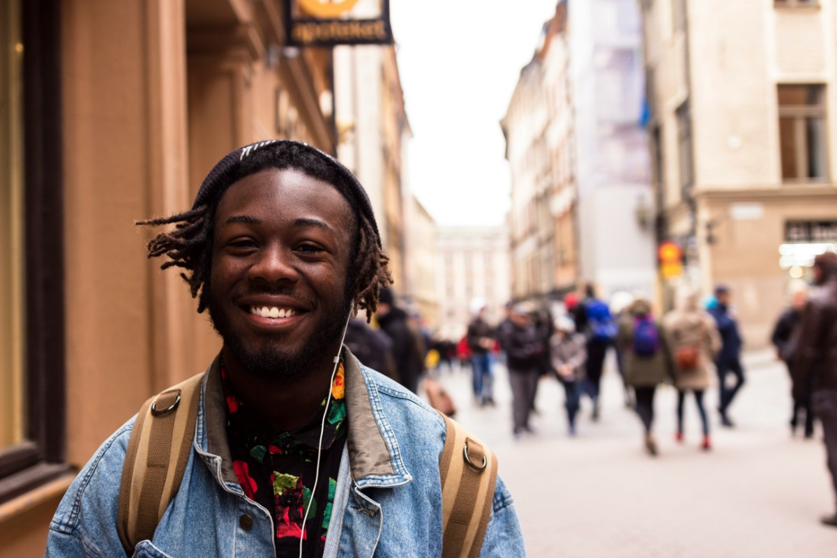 A person with a denim jacket smiles while standing on a busy street lined with buildings. People walk in the background.