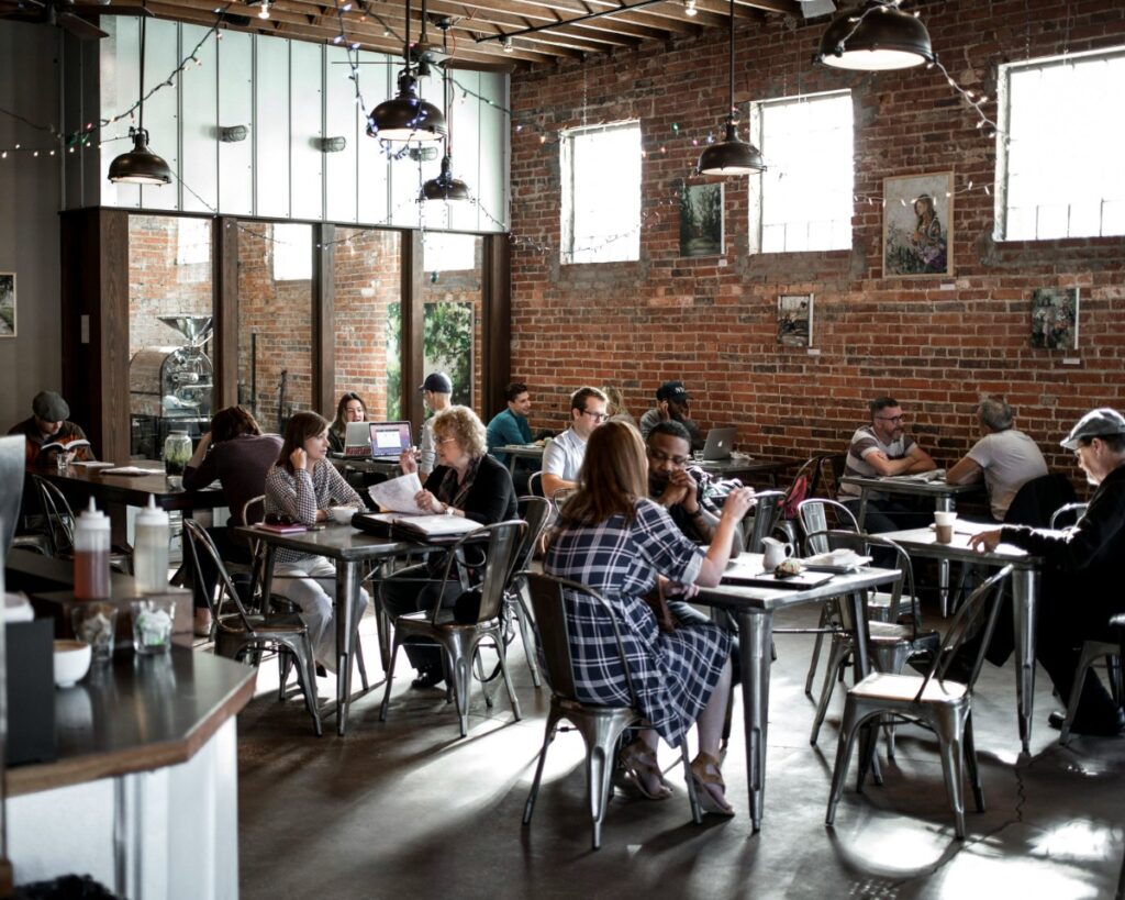 People sitting at tables inside a cozy cafe with brick walls and hanging lights. Some are using laptops, while others are talking or dining.